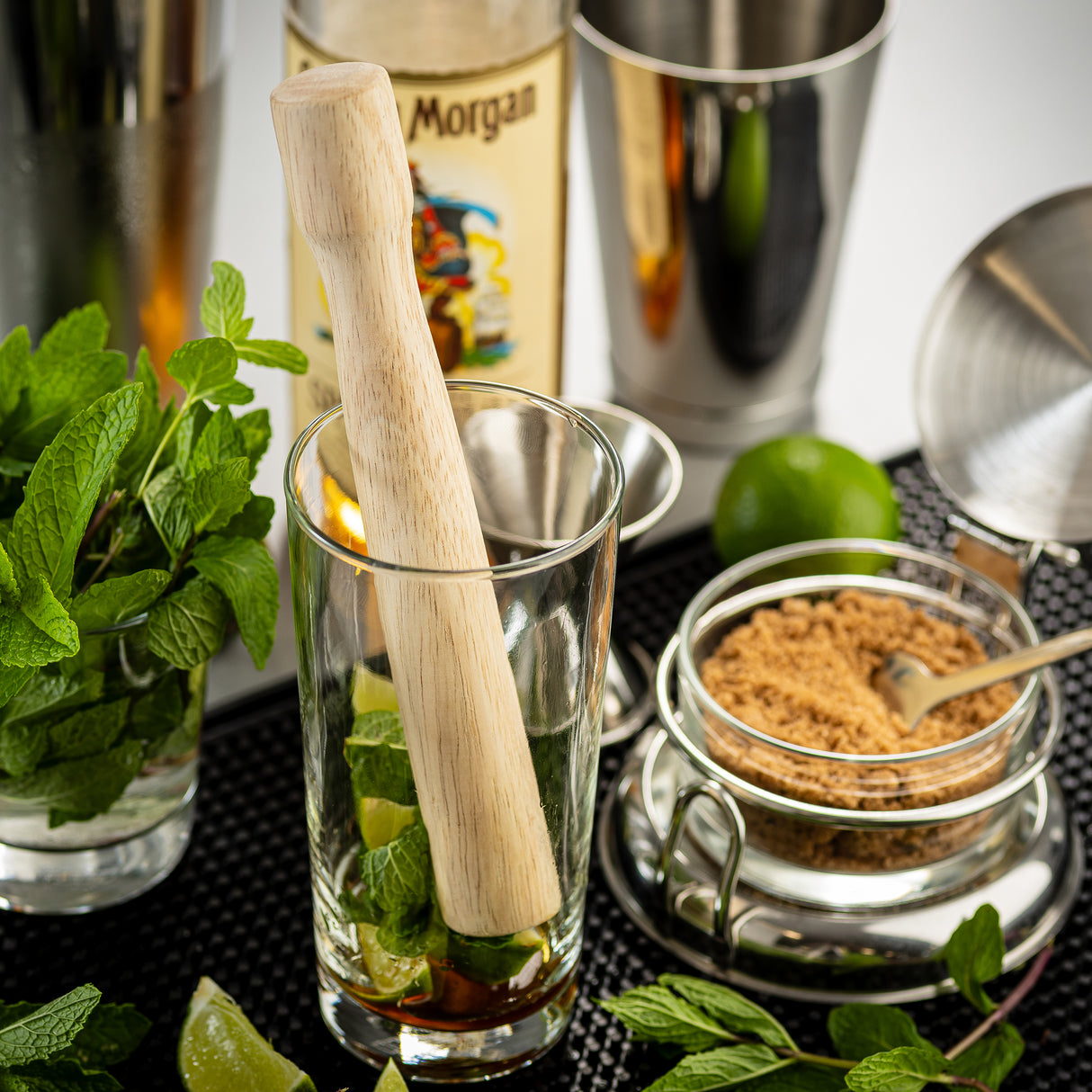 A TableCraft muddler made of natural wood rests in a glass filled with lime wedges, mint leaves, and brown sugar. Fresh mint, a lime, and a jar of brown sugar are nearby, with rum and metal cocktail tools set on a bar mat in the background.