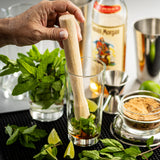 A hand uses a TableCraft natural wood muddler to crush fresh mint leaves in a glass. Surrounding the glass are lime wedges, a bowl of brown sugar, mint sprigs, Captain Morgan rum, a metal jigger, and bar tools on a black mat.