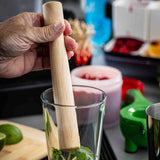 A hand uses a natural wood TableCraft muddler to crush mint leaves in a glass. In the background, cut limes, a green citrus squeezer, metal shaker, and colorful ingredients hint at cocktail preparation.