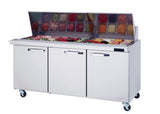 A commercial sandwich prep table with three lower cabinets and a glass-covered mega top prep section displays fresh ingredients like vegetables, deli meats, and cheese in organized containers, all set on wheels for easy mobility.