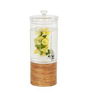 A clear glass beverage dispenser with an infusion chamber, lid, and spigot is filled with water and slices of lemon and lime. The dispenser sits on a round wood base, all set against a plain white background.