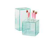 Two clear, textured glass blocks are shown; one taller and rectangular, the other shorter and square. The square block, reminiscent of a Glacier Display Riser from Cal-Mil, holds three pink drinks in shot glasses, each topped with a strawberry. White background.