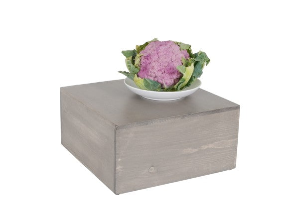 A head of purple cauliflower with green leaves sits on a white plate, which is placed on top of a large Cal-Mil Aspen Riser made from gray-washed pine wood, set against a plain white background.