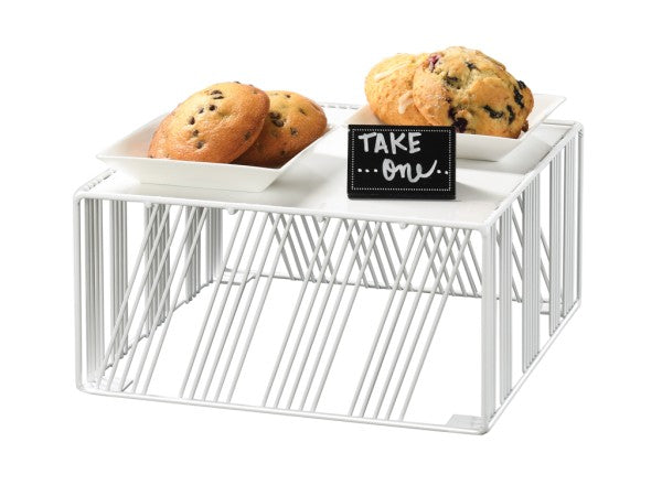 A white Portland Riser display stand holds two square white plates with chocolate chip cookies and muffins. In front of the plates sits a small blackboard sign with white handwritten text reading TAKE ONE. The background is plain white.