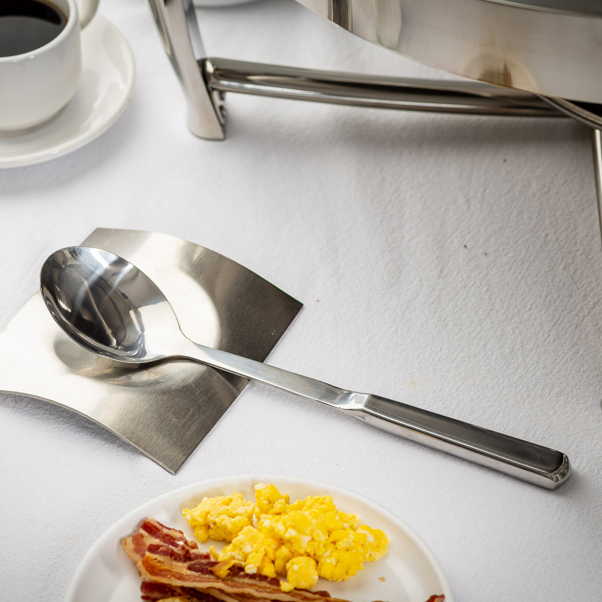 A large dishwasher safe serving spoon rests on a stainless steel plate atop a white tablecloth. Nearby are a cup of coffee, part of a chafing dish, and a plate with scrambled eggs and bacon strips.
