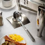 A large stainless steel serving spoon rests on a square metal plate atop a white tablecloth. Nearby are a plate with scrambled eggs, bacon, and sausage, a cup and saucer, a pepper shaker, and a shiny chafing dish, all arranged neatly.