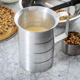 A 2 qt aluminum measuring cup filled with dry cornmeal sits on a marble countertop. In the background, there's a pie topped with nuts, a small metal cup of walnuts, and a stainless steel mixing bowl with a black spatula inside.