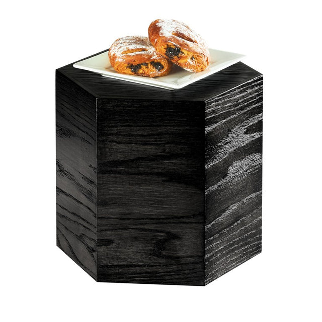 A hexagonal black oak wood table with visible grain patterns holds a square white plate with two powdered pastries filled with a dark filling, set against a plain white background.