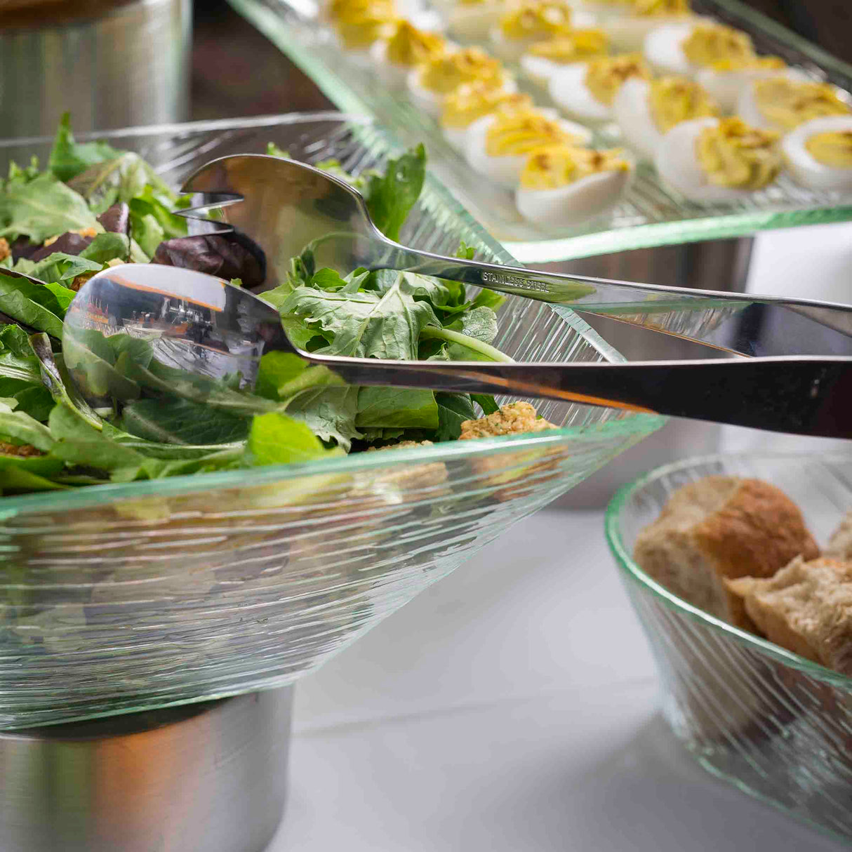 A glass bowl of fresh mixed greens with stainless steel serving tongs rests on a buffet table. In the background, deviled eggs and a bowl of sliced bread are arranged in clear glass dishes on a white tablecloth.