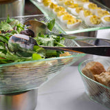 A glass bowl of fresh mixed greens with stainless steel serving tongs rests on a buffet table. In the background, deviled eggs and a bowl of sliced bread are arranged in clear glass dishes on a white tablecloth.