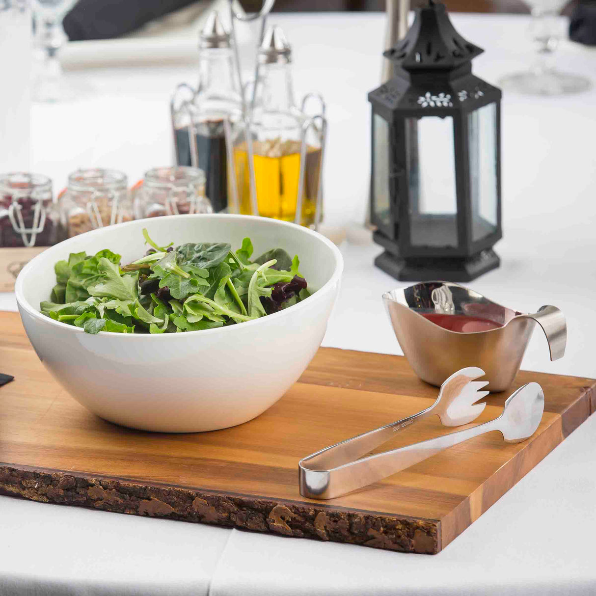 A white bowl of mixed leafy greens sits on a wooden board with stainless steel serving tongs. Beside it is a small metal gravy boat with red dressing. In the background are glass bottles of oil, spice jars, and a black lantern on a white tablecloth.