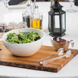 A white bowl of mixed leafy greens sits on a wooden board with stainless steel serving tongs. Beside it is a small metal gravy boat with red dressing. In the background are glass bottles of oil, spice jars, and a black lantern on a white tablecloth.