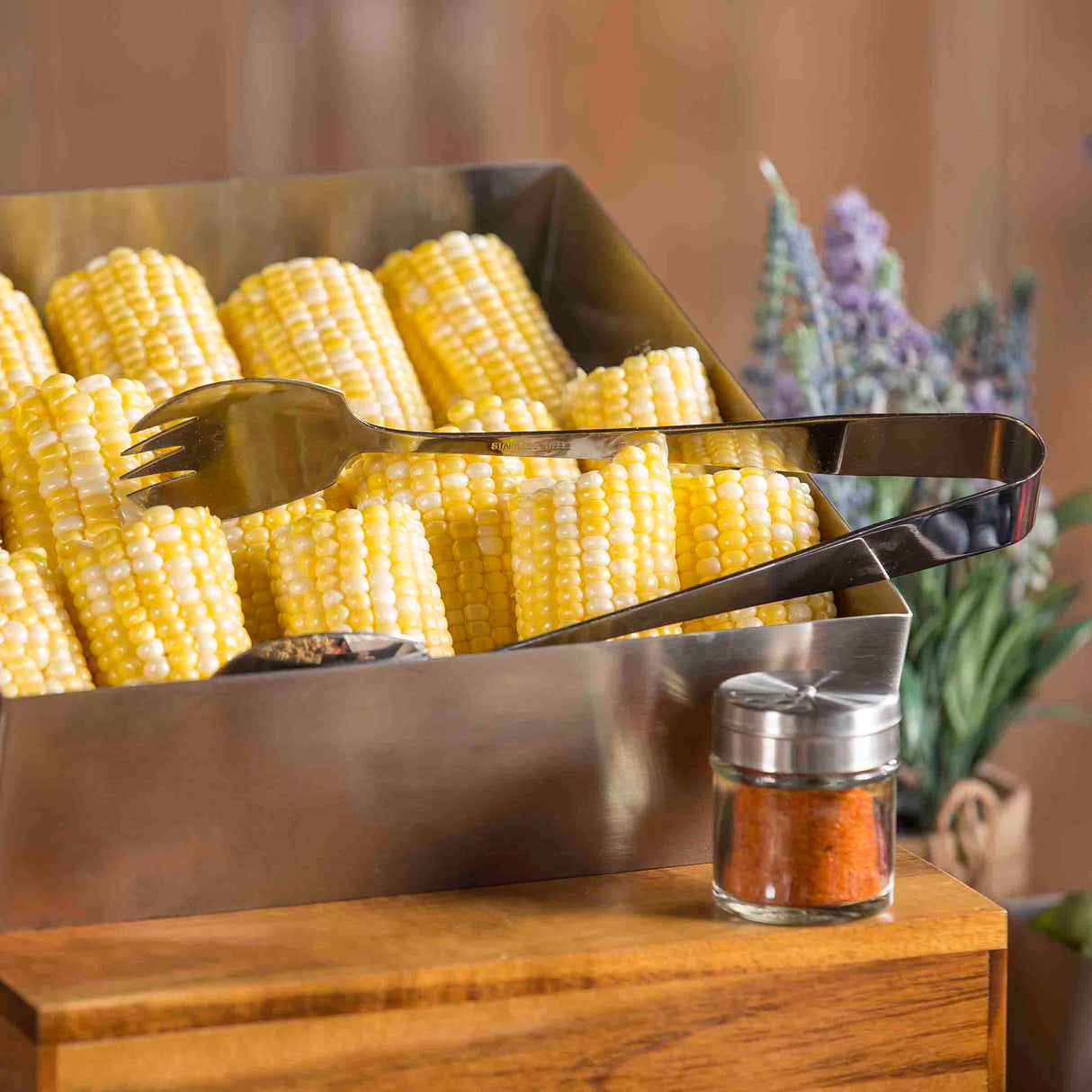 A metal tray filled with yellow and white corn on the cob is shown, with stainless steel serving tongs resting on top. In the foreground, a small glass jar of red seasoning sits, while blurred lavender flowers appear in the background.