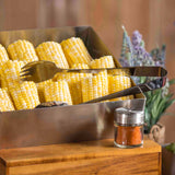 A metal tray filled with yellow and white corn on the cob is shown, with stainless steel serving tongs resting on top. In the foreground, a small glass jar of red seasoning sits, while blurred lavender flowers appear in the background.