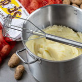 A metal pot filled with mashed potatoes sits on a countertop. A dishwasher safe potato masher with a stainless steel handle rests on the mash. Nearby are whole russet potatoes, a red mesh bag, and a white box labeled potatoes.