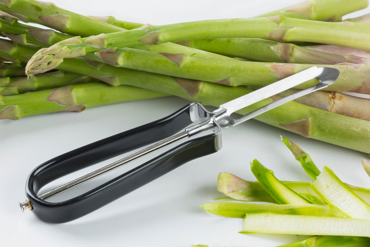 A black-handled vegetable peeler with a stainless steel blade lies on a white surface, surrounded by fresh green asparagus spears. Some asparagus tips are whole, others partially peeled, with green peelings scattered nearby.