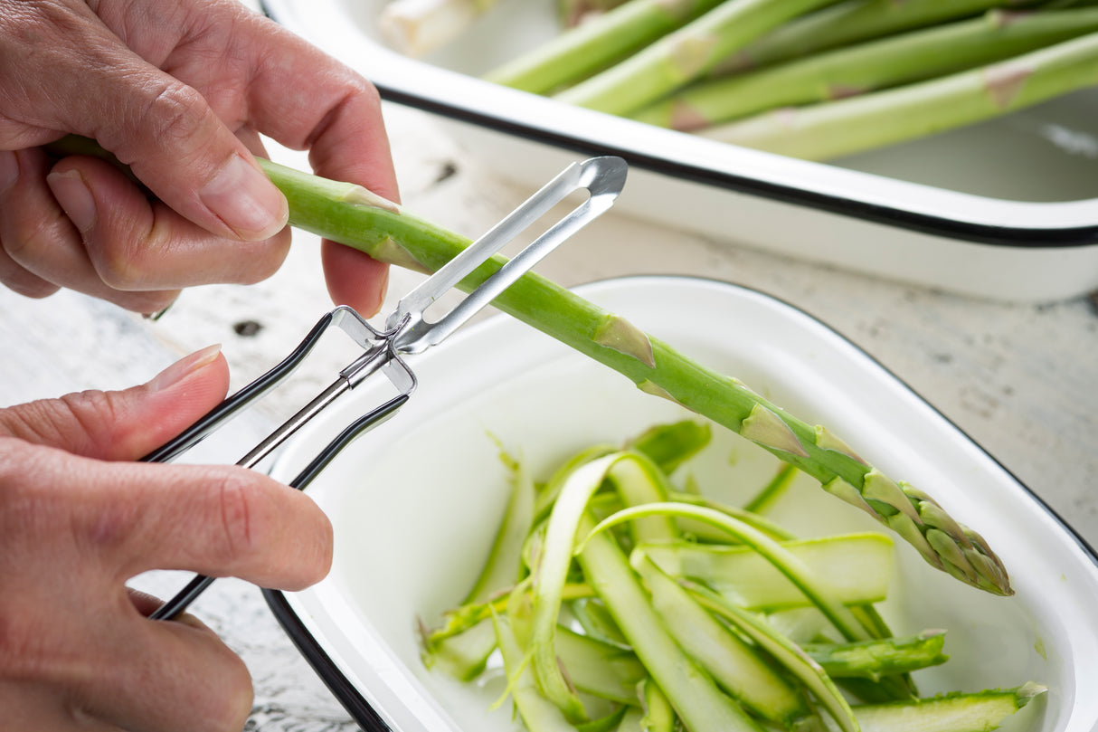 Close-up of hands peeling a fresh green asparagus spear with a peeler featuring a dishwasher safe stainless steel blade. A white dish underneath collects the thin green asparagus peels, while more unpeeled spears are visible in the background.
