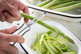 Close-up of hands peeling a fresh green asparagus spear with a peeler featuring a dishwasher safe stainless steel blade. A white dish underneath collects the thin green asparagus peels, while more unpeeled spears are visible in the background.