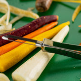 A close-up of colorful carrots—white, yellow, orange, and purple—on a green surface, with an Economy Peeler in the foreground. Vegetable peels are scattered around, showing the carrots being prepared with this dishwasher safe tool.