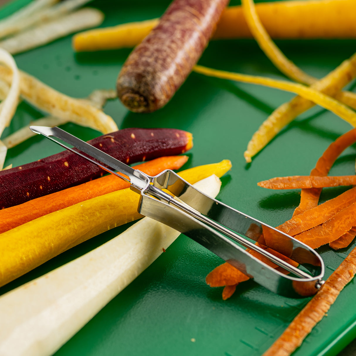 A tempered steel Economy Peeler rests on a green surface surrounded by colorful carrot peels and whole carrots in purple, yellow, and orange. Peeled strips and fragments scattered around highlight convenient, dishwasher safe food preparation.