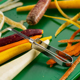 A tempered steel Economy Peeler rests on a green surface surrounded by colorful carrot peels and whole carrots in purple, yellow, and orange. Peeled strips and fragments scattered around highlight convenient, dishwasher safe food preparation.