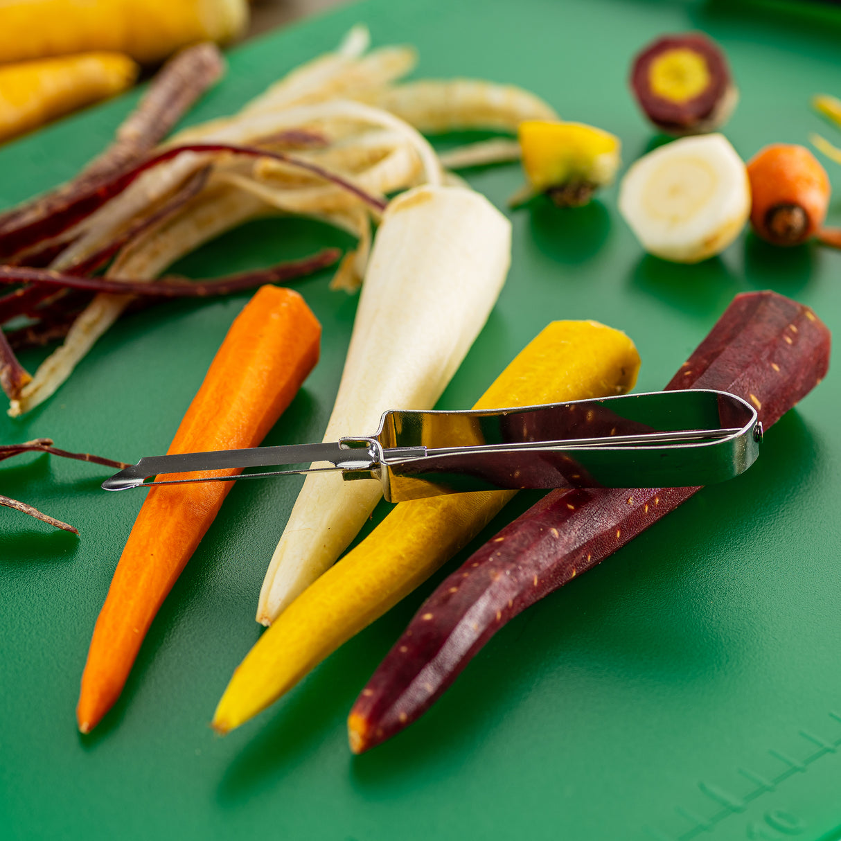 An Economy Peeler with a tempered steel blade rests on colorful peeled carrots—orange, yellow, white, and purple—on a green cutting board. Peeled carrot skins and carrot tops are scattered around, with a sliced purple carrot in the background.