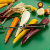 An Economy Peeler with a tempered steel blade rests on colorful peeled carrots—orange, yellow, white, and purple—on a green cutting board. Peeled carrot skins and carrot tops are scattered around, with a sliced purple carrot in the background.