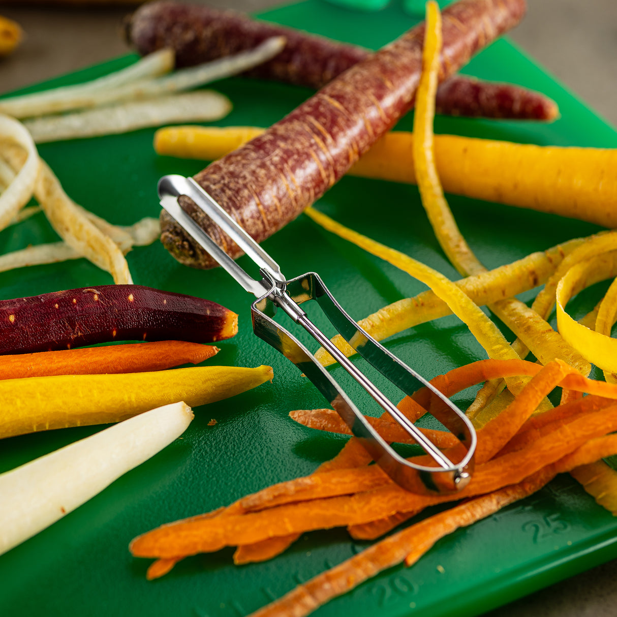 A tempered steel Economy Peeler rests on a green cutting board surrounded by colorful carrot varieties—orange, yellow, purple, and white—both whole and peeled with scattered carrot peels. The peeler is dishwasher safe, making meal prep quick and convenient.