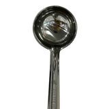 A shiny, dishwasher safe stainless steel measuring spoon with a round bowl and a long handle is shown on a white background. The reflective surface of the spoon displays some distorted reflections.