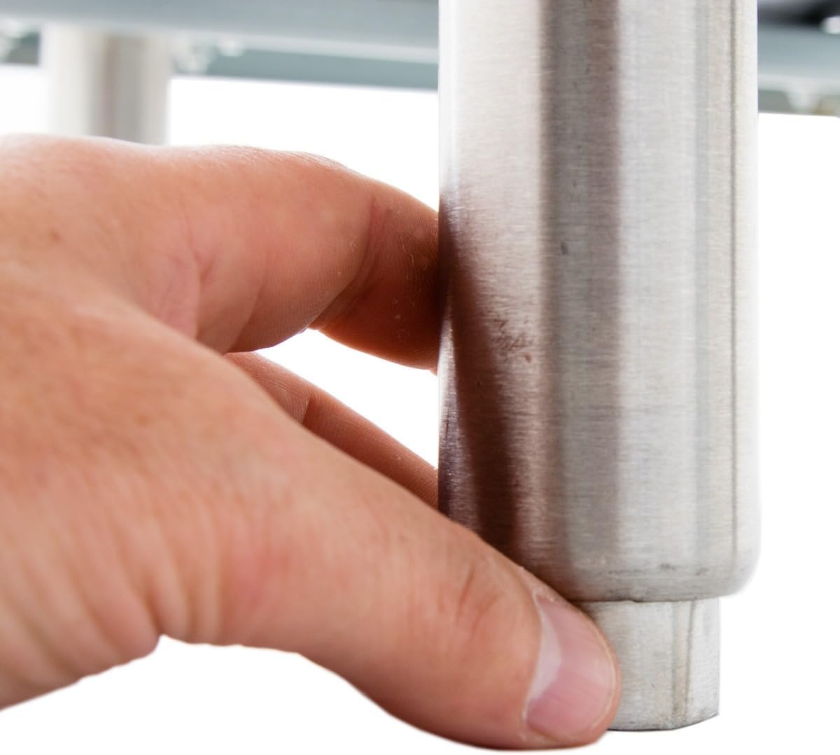 A close-up of a persons left hand holding the bottom of a round, metallic table leg—possibly part of a countertop griddle—with a smooth surface. The hand grasps the leg firmly, set against a white background with part of the tabletop faintly visible above.