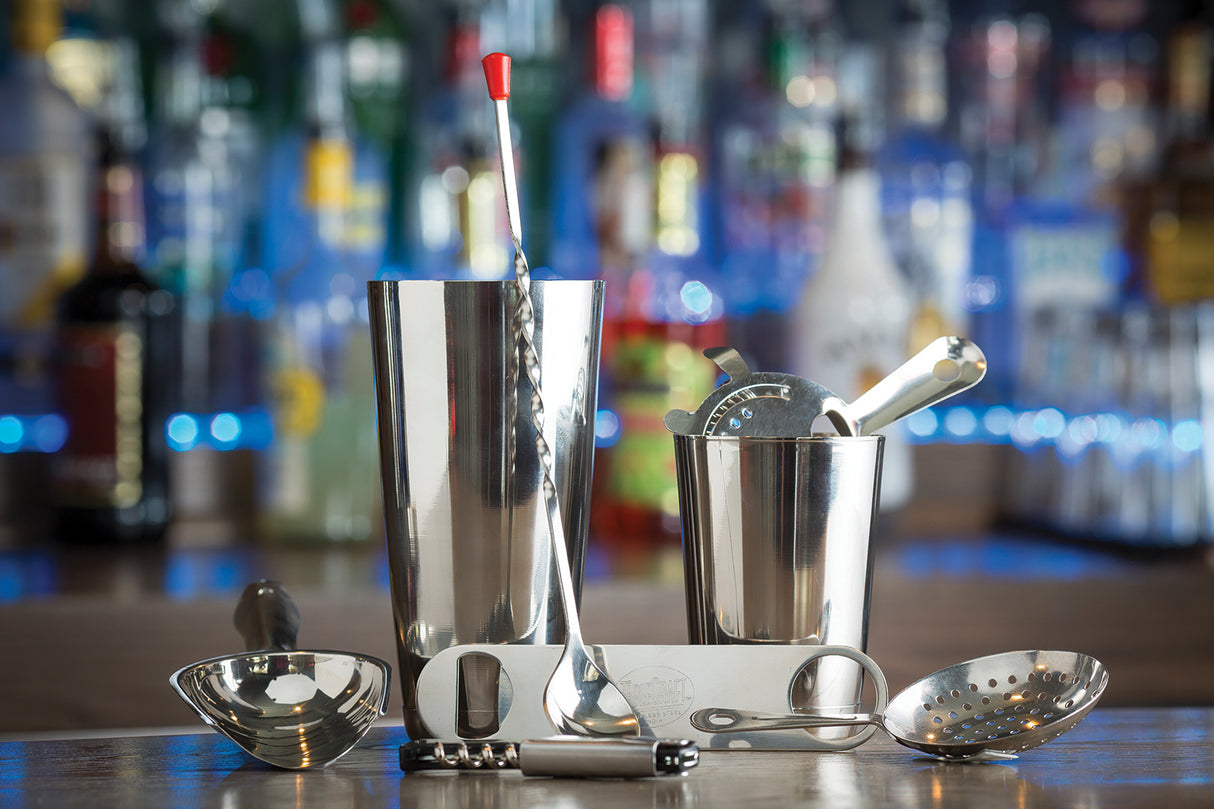 A set of shiny stainless steel bar tools—including a shaker, jigger, strainer, bar spoon, tongs, corkscrew, and muddler—are arranged on a bar counter. Bottles of liquor blur in the colorful background, suggesting a lively bar setting.