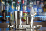 A set of shiny stainless steel bar tools—including a shaker, jigger, strainer, bar spoon, tongs, corkscrew, and muddler—are arranged on a bar counter. Bottles of liquor blur in the colorful background, suggesting a lively bar setting.