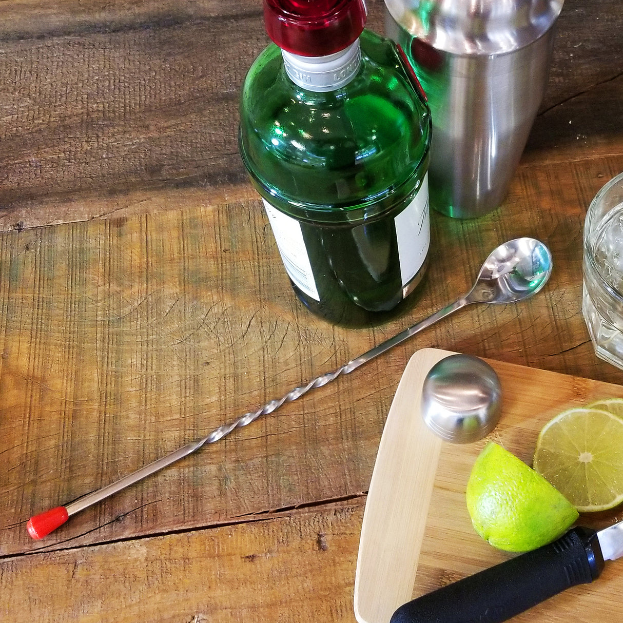 A 12 inch stainless steel bar spoon with a red tip lies on a wooden table near a green bottle, metal cocktail shaker, glass, cutting board with sliced lime, and bottle cap. The scene suggests cocktail preparation.