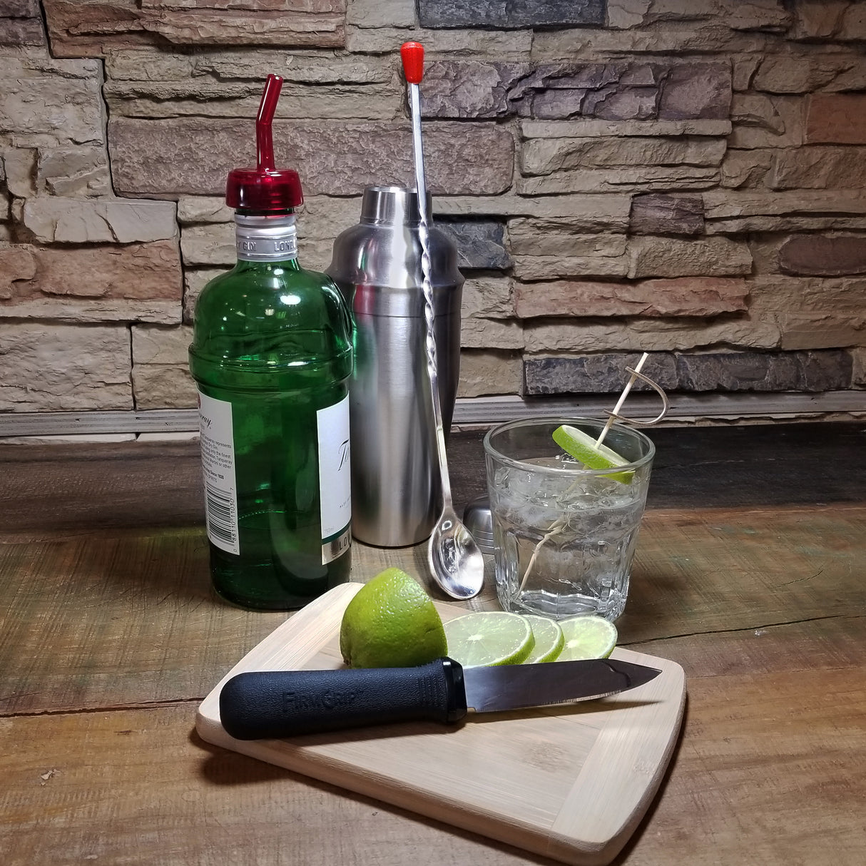 A bottle of gin, a stainless steel bar spoon, a metal shaker, and a glass of gin and tonic with lime sit on a rustic wooden surface. In front, a cutting board holds a knife, a halved lime, and three lime slices. The background is a stone wall.