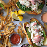 Two lobster rolls on toasted buns with lettuce and creamy filling, served with French fries, lemon wedges in a stainless steel sauce cup, ketchup, coleslaw, and a cup of pink dipping sauce—all arranged on a white surface.