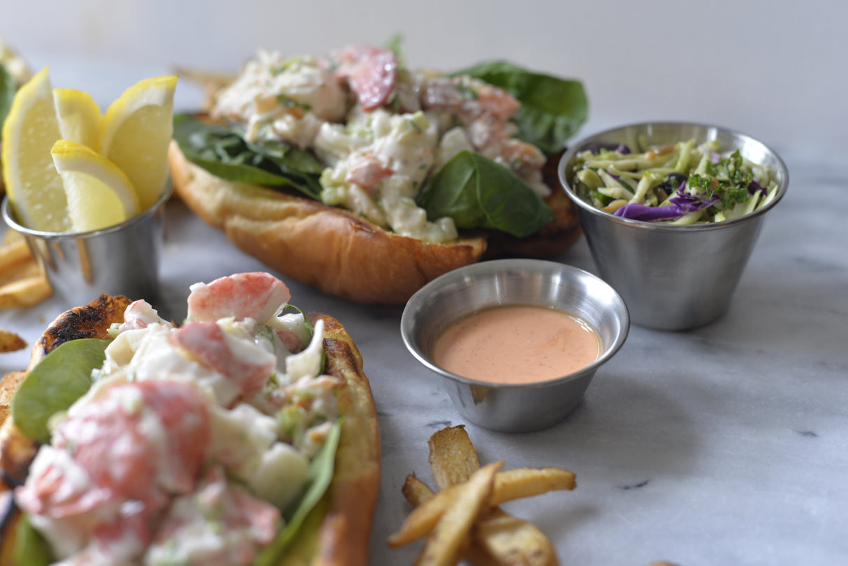 Two lobster rolls on toasted buns with leafy greens and creamy filling are served with fries, pickle wedges, a cup of coleslaw, and a 4 oz stainless steel sauce cup filled with pink dipping sauce on a light marble surface.