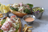 Two lobster rolls on toasted buns with leafy greens and creamy filling are served with fries, pickle wedges, a cup of coleslaw, and a 4 oz stainless steel sauce cup filled with pink dipping sauce on a light marble surface.
