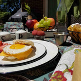 A close-up of a breakfast table outdoors shows a plate with a sunny-side-up egg, cheese, biscuit, and a stainless steel sauce cup. Nearby are two cups, fruit baskets, a glass bottle, and a grill in the background atop a floral tablecloth.