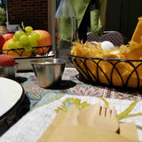 A close-up of a table set outdoors with a plate, wooden cutlery, and napkin in the foreground. A stainless steel sauce cup sits beside a basket of yellow fabric with two eggs, fruit bowl, metal cup, and glass bottle. Green leaves and brick wall behind.