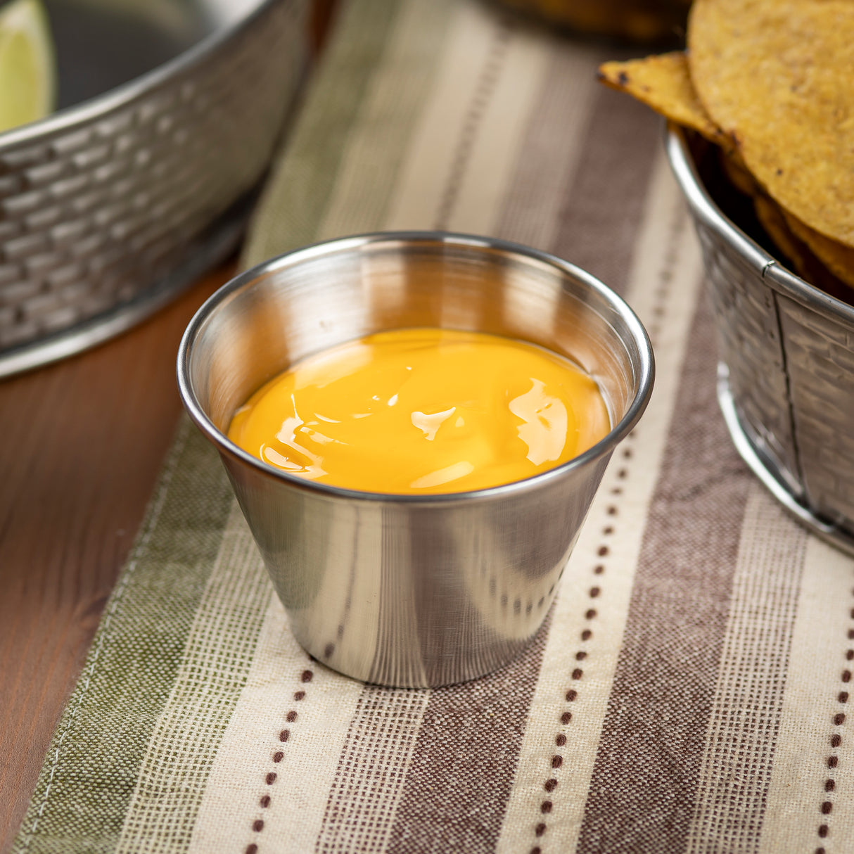 A 2 oz stainless steel sauce cup filled with creamy orange cheese sauce sits on a striped fabric placemat, next to a metal basket of tortilla chips. The scene suggests a snack or appetizer setup, with soft, natural lighting.