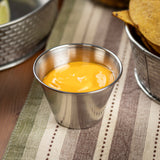 A 2 oz stainless steel sauce cup filled with creamy orange cheese sauce sits on a striped fabric placemat, next to a metal basket of tortilla chips. The scene suggests a snack or appetizer setup, with soft, natural lighting.
