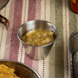 A 2 oz stainless steel sauce cup filled with chunky green salsa sits on a striped cloth surface. Visible veggie bits enhance its appeal. Part of a metal basket with tortilla chips appears in the lower left, with another basket partially seen on the right.