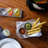 A stainless steel basket lined with white paper holds four taquitos, a small sauce cup of salsa, and another of guacamole. Nearby are two drinks with salt rims and lime wedges, plus a plate and a napkin, all on a wooden table.