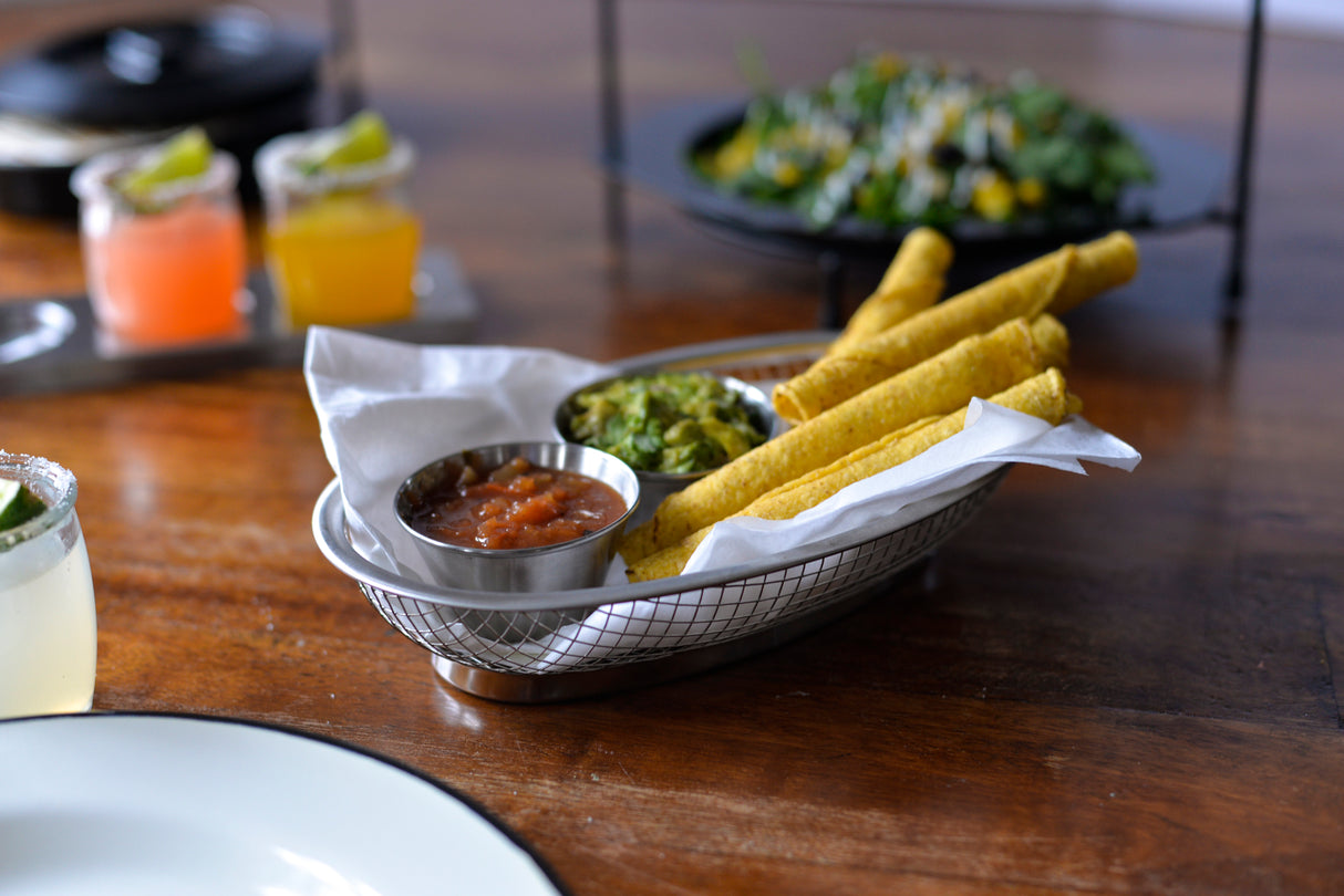 A stainless steel wire basket lined with paper holds four rolled taquitos, a small sauce cup of salsa, and a cup of guacamole. In the background are drinks with lime wedges and a plate of salad with corn on a wooden table.