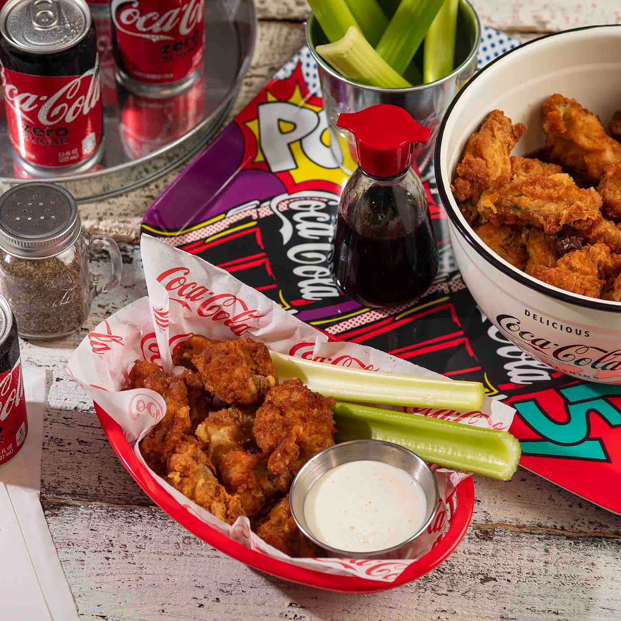 A basket of fried chicken, celery sticks, and ranch dip sits on a table beside a stainless steel sauce cup, cans of Coca-Cola Zero Sugar, a soy sauce dispenser, and colorful comic book covers as placemats.
