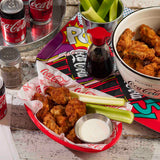 A basket of fried chicken, celery sticks, and ranch dip sits on a table beside a stainless steel sauce cup, cans of Coca-Cola Zero Sugar, a soy sauce dispenser, and colorful comic book covers as placemats.