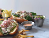 A lobster roll with chunks of lobster, spinach, and creamy dressing sits on a toasted bun. Sides include lemon wedges, thin fries, a 6 oz stainless steel sauce cup, and coleslaw with green and purple cabbage on a marble surface.