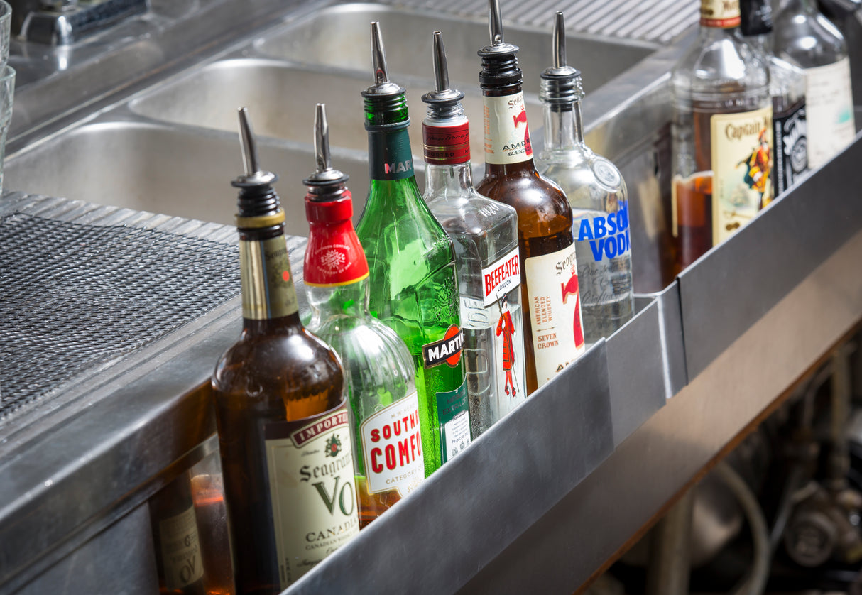 A row of assorted liquor bottles with pour spouts is lined up on an adjustable stainless steel speed rail near a metal sink. Bottles include vodka, gin, vermouth, and other spirits, some with visible labels and bar equipment in the background.