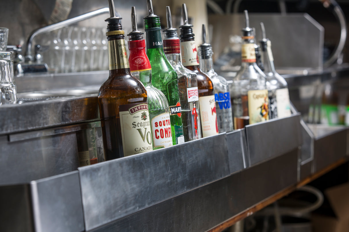 A row of assorted liquor bottles with pour spouts sits on an adjustable stainless steel speed rail that holds 8 bottles. Empty glasses are stacked in the background near a sink, indicating a bar or restaurant setting.