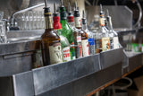 A row of assorted liquor bottles with pour spouts sits on an adjustable stainless steel speed rail that holds 8 bottles. Empty glasses are stacked in the background near a sink, indicating a bar or restaurant setting.